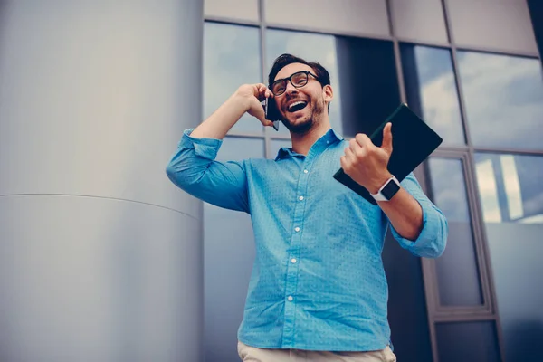 Emotional male student in eyewear excited with finishing work on educational project sharing good news with friends during mobile conversation, entrepreneur happy getting offer for startup project