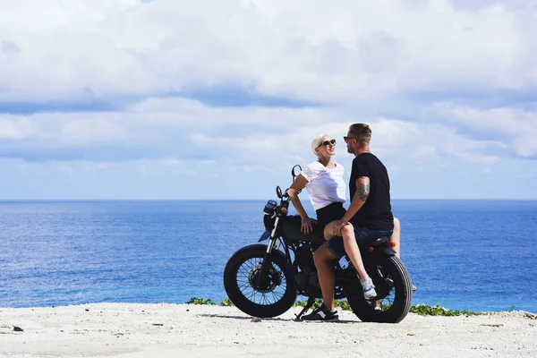 Romantic couple in love spending time on sea shore sitting on motorcycle enjoying being together, young male and female bikers having riding tour on cost spending summertime weekends near ocean