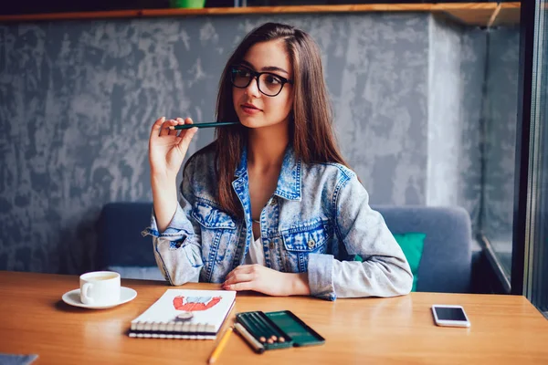 Elinde kalem tutan ve uzak çizimler için Not Defteri ile masaüstünde otururken seyir şık gözlük çekici profesyonel tasarımcısında. Güzel ve genç birbayan çizim için yeni fikirler üzerinde düşünme