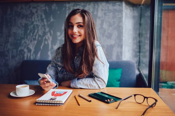 Neşeli genç kadın arkadaşlarıyla sohbet fincan lezzetli kahve ve tasarım için Not Defteri ile masada oturan cep telefonunda mesajlaşma yarım uzunluk portre rahat kafeteryada kapalı skeçler