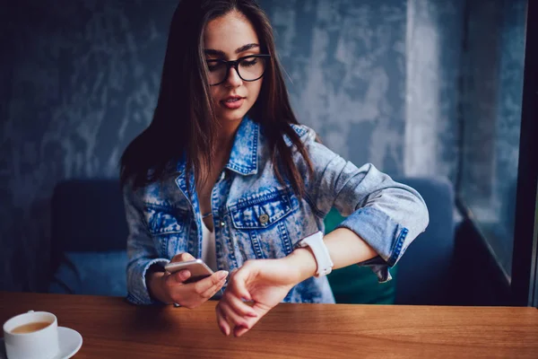 Busy female manager in stylish eyeglasses looking at watch while holding telephone in hand and hurry on meeting.Businesswoman keeping track of time sitting at table in coffee shop interior