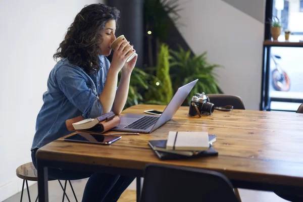 Stylish hipster girl drinking tasty coffee while studying at modern netbook computer connecting to wireless internet sitting at wooden table with vintage camera and books in coworking space