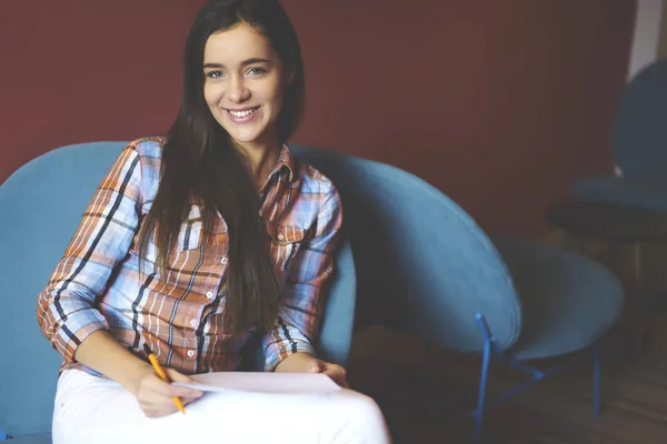 Portrait of charming female student sitting in cozy chair in university campus enjoying doing creative home task.Clever young woman in casual wear pondering on planning project looking at camera