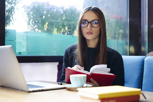 Yarım uzunluk portre zeki öğrenci ders kitabı elinde tutan ve kamera için 4 g bağlanan dizüstü bilgisayar ile oturan bakarken not yazmak daha iyi görüş için trendy gözlük içinde Internet