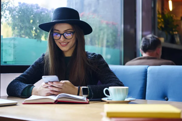 眼鏡と一杯のコーヒーとお気に入りの本屋内カフェテリアで余暇を過ごすの携帯電話で友人から面白い Sms メッセージを読んでおしゃれな帽子でスタイリッシュな内気な少女の笑顔 — ストック写真