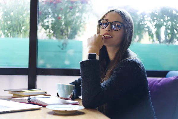 Portrait of brunette teen hipster girl in eyewear enjoying free time in cafe interior looking at camera. Attractive young woman in casual wear drinking coffee while resting doing coffee work