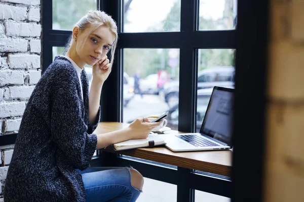 Portrait of pensive blonde female copywriter earning money online creating multimedia content while sitting in cafe interior,thoughtful freelancer waiting for feedback from banking service using phone