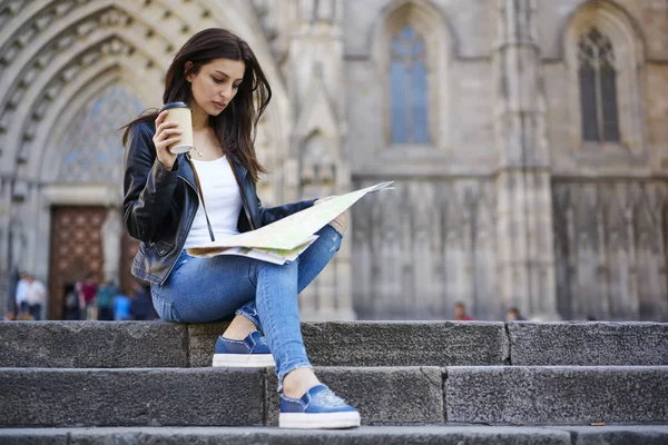 Concentrated young tourist dressed in trendy casual wear holding map deciding where to go in ancient city, charming hipster girl in trendy apparel sitting with coffee to go searching right direction