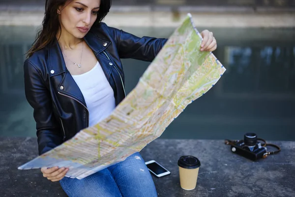 Concentrated young woman reading map creating city tour way while recreating in weekend trip, attractive female traveler searching right destination to show places in town sitting on urban settings