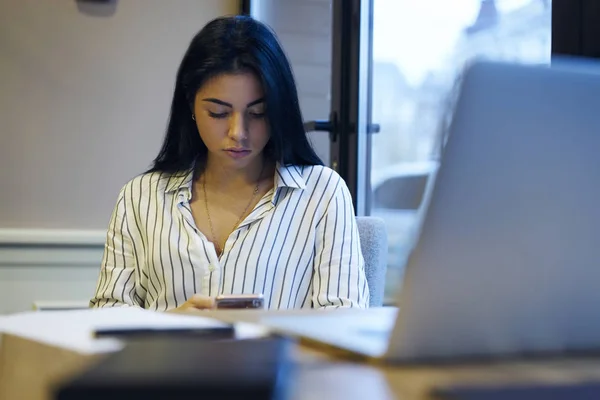 オフィス モバイル更新アプリケーション オンラインの前に座ってデスクトップで設定を行う物思いにふける若い女性マネージャーで作業しながらスマート フォンでブルネットのビジネス女性読書メッセージを集中 — ストック写真