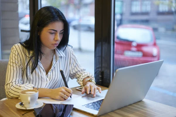 Pensive young female economist browsing information on laptop computer using application for checking accountings,creative web designer working on freelance using modern technology in coffee shop