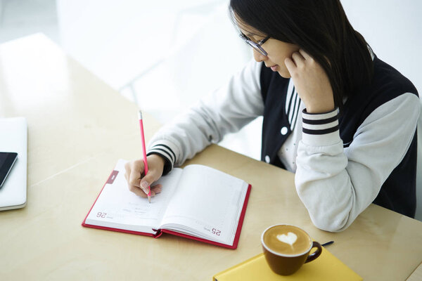 Pensive brunette teen pupil in eyewear concentrated on making homework sitting at desktop in library, clever international student writing report in notebook analyzing information in library