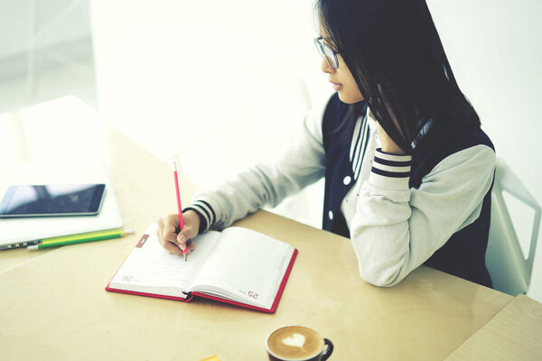 Skilled female trainee concentrated on writing publication noting ideas with pen sitting in coworking space near copy space for advertising, clever teen pupil doing homework pondering on task