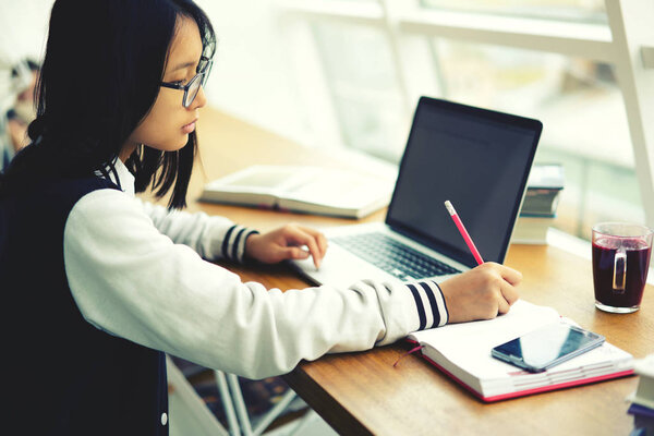 Clever international student concentrated on writing in notebook preparing homework in coworking space, young pupil in eyewear making researches via laptop analyzing information from network 