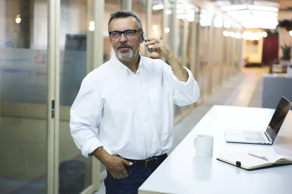 Professional lawyer in formal clothing talking on mobile telephone discussing important questions while standing in office.Businessman in spectacles having conversation on mobile booking tickets online