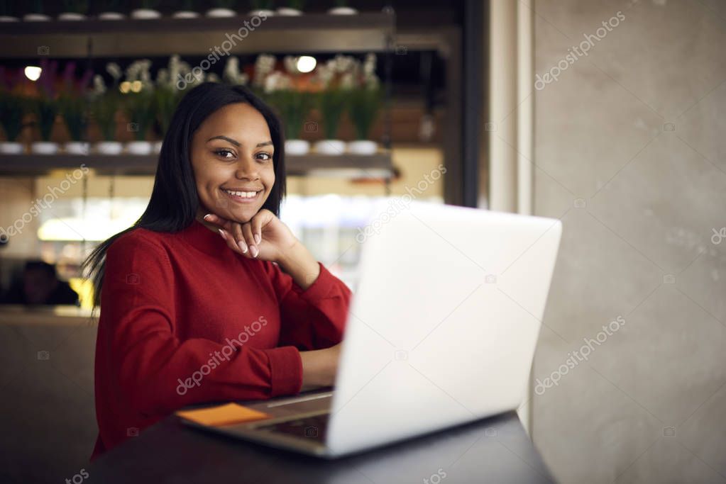 Portrait of afro american student sitting in coffee shop on break looking at camera, charming dark skinned hipster girl spending leisure in cafe using laptop computer for blogging in network