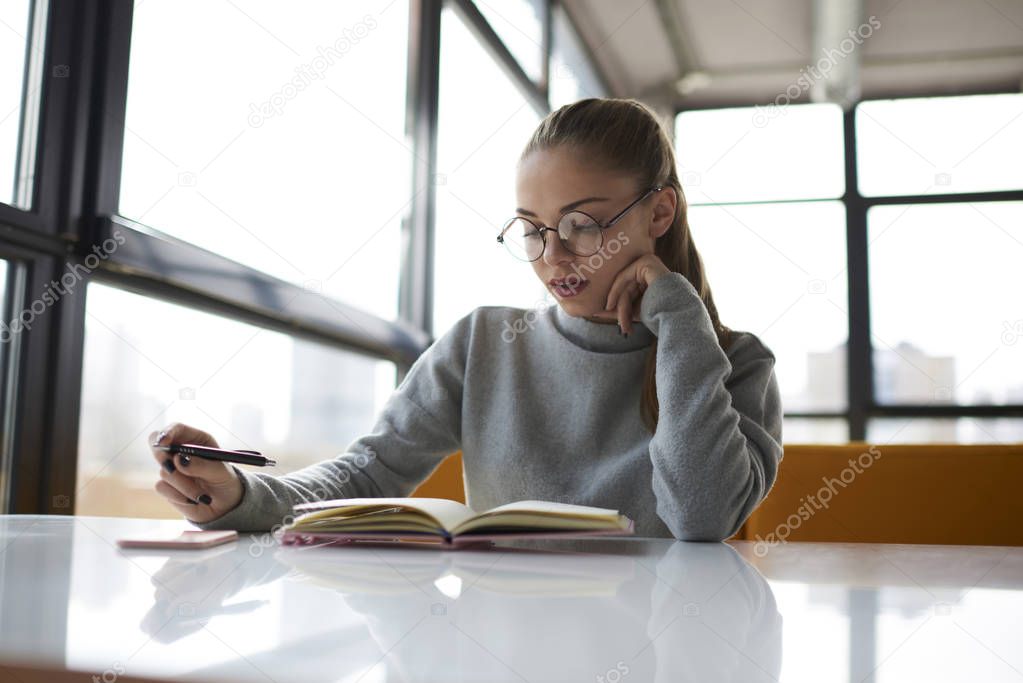 Pensive young student in stylish eyewear reading book studying in modern interior library.Thoughtful attractive female manager checking planning and numbers for startup project written in notepa