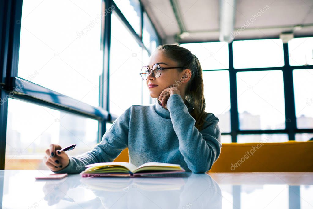 Beautiful female student in eyeglasses looking away in window sitting in cafe while doing homework tasks dreaming about vacations.Thoughtful business woman making plan for project enjoying leisure