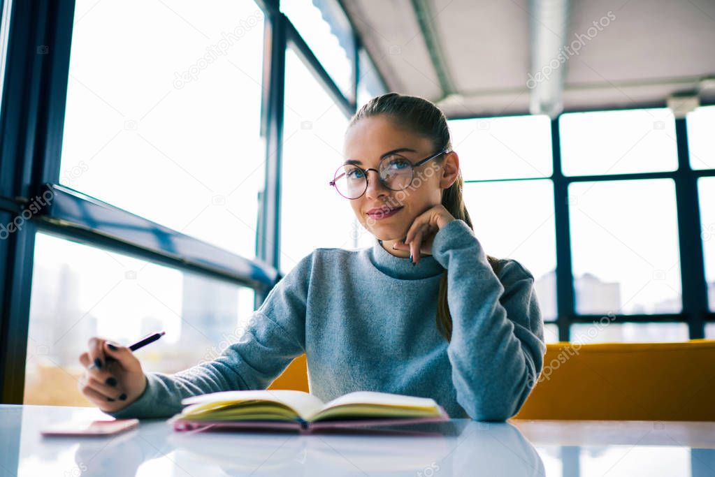 Portrait of beautiful young student in eyeglasses studying in modern interior library preparing for university examinations.Confident businesswoman creating plan for startup project making notes