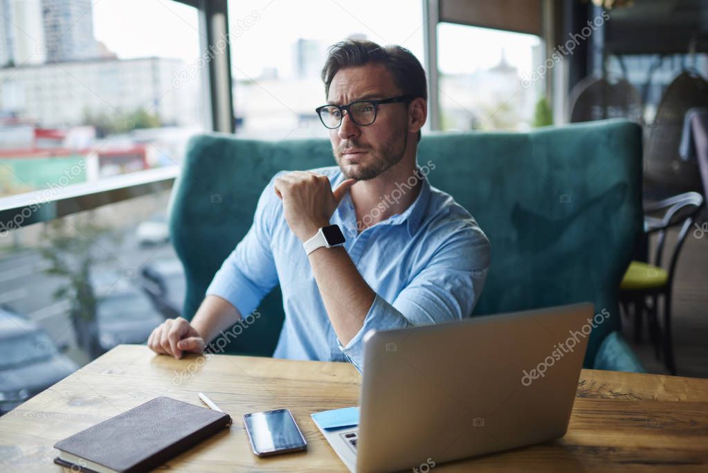 Pensive owner f business corporation thinking about new strategy while sitting in cafe interior with laptop, handsome manger in eyeglasses concentrated while creating planning for startup projec