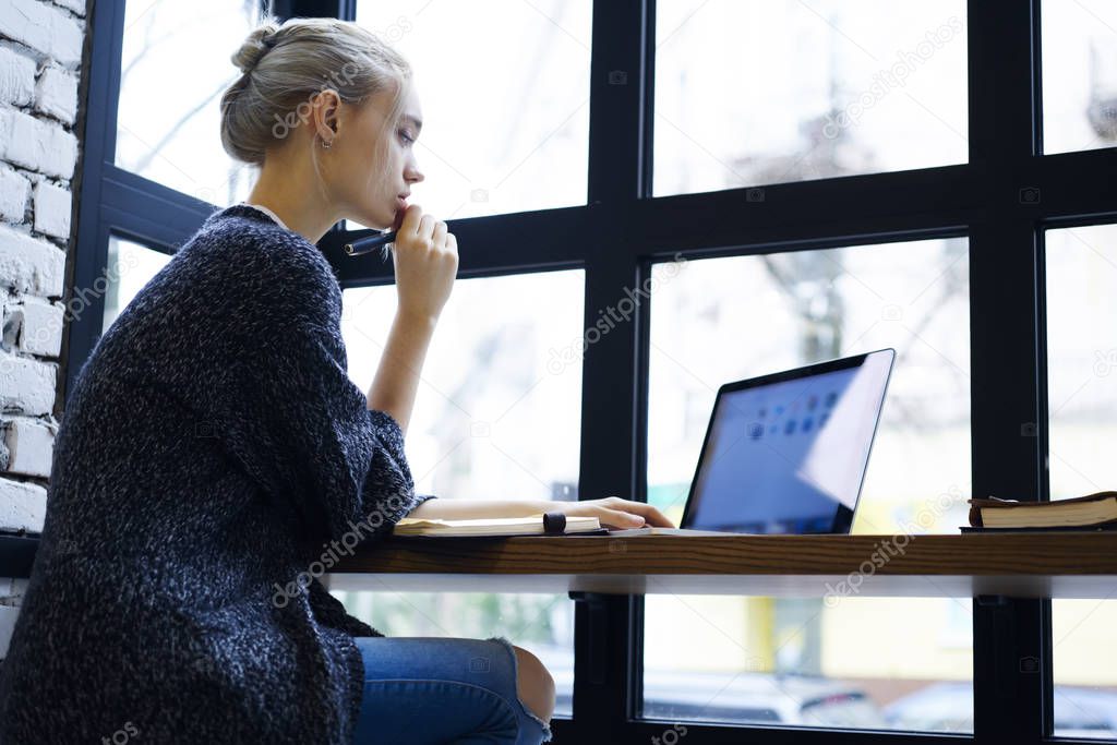 Charming pensive female web designer working on freelance concentrated on modeling project using application on laptop computer with mock up screen earning money online sitting in cafe interior