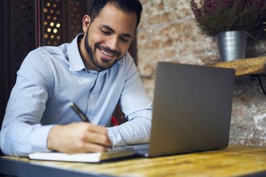 Neşeli yakışıklı erkek tasarımcı çizim kroki üzerinde dizüstü bilgisayar uygulaması, kafede oturan başlangıç planlama iç yapım genç profesyonel iş Yöneticisi modelleme önce not defteri
