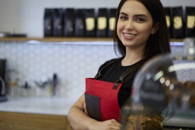 Kameraya bakarak ve gülümseyen siyah saçlı neşeli garson yarım uzunluk portre. Çekici olumlu genç kadın çalışma barista şık kahve dükkanında. Kısa mesaj reklam için yer alan kopyalama