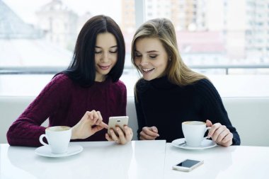 İki gülümseyen arkadaş komik sohbet restoranda lezzetli kahve ile oturan modern telefon üzerinde okuma. Hippi kız rekreasyon vaktinizi sıcak içecekler ile kafede ve üzerinde hücresel mesajlaşma