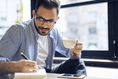 Dalgın yakışıklı raslantı giyimli grafik tasarımcı kafeteryasında Kupası Kahve çizim planı defter, genç öğrenci rapor yazma üzerinde konsantre fikirlerin belirterek gözlük zevk çalışma