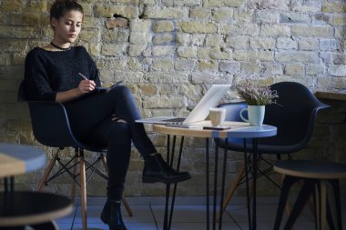 Konsantre öğrenci dizüstü dizüstü bilgisayar aygıt kapalı şık kafeteryasında oturan Eskizler çizmeye gündelik kıyafeti giymiş. Not yazmak ve Planlama Teşkilatı içinde not etmek dalgın genç kadın  