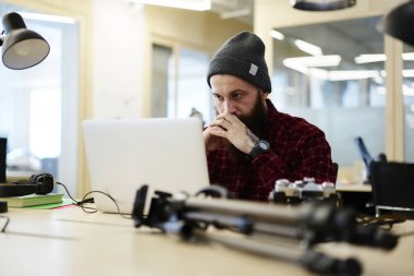 Ciddi yetenekli fotoğrafçı dikkatlice dizüstü kablosuz internet'e bağlı video izlerken trendy kıyafeti giymiş. Genç modern, çalışma alanı'nda oturan aygıt üzerinde çalışan konsantre