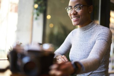 Neşeli kadın siyah modeli almak Eyewear cephe süsleme hobi, yaratıcı iş bulanık reklam alan üzerine oturan ABD'li fotoğrafçı memnun afro zaman harcama üzerinde vintage fotoğraf makinesi bulanık
