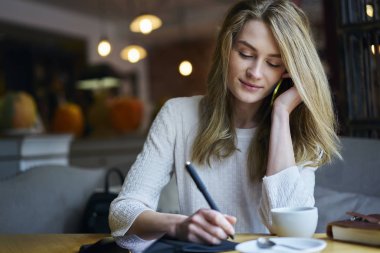 Kahve masasi coworking oturan Not Defteri'nde metin bilgileri yazılırken cep telefonu ile konuşurken güzel meşgul kadın Yöneticisi. Kopyalama alanı alanı reklam metin iletinizi için