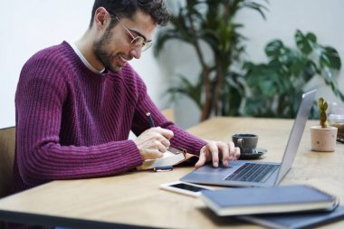 Gülümseyen dizüstü Web sitelerinde bilgi arama ve genel fikirler Kayıt Defteri'nde gözlük girişimci sakallı. Pozitif genç serbest meslek sahibi uzaktan kapalı modern aygıtıyla çalışma