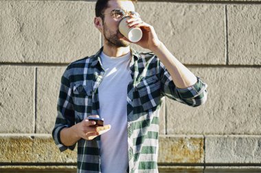 Genç yakışıklı gezgin gri taş duvarın karşısında dururken lezzetli kahve zevk trendy kıyafetler giymiş. Rekreasyon vakit aroma içecek ve gadget ile gözlük içinde erkek blogger
