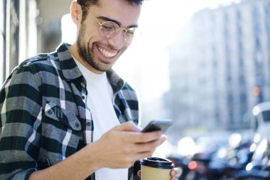 Neşeli mutlu blogger manifatura komik geribildirim Web sitesinde kullanarak yüksek hızda Lte 4 g smartphone içinde güneşli gün sokakta geziniyor ise internet. Hipster adam okuma mesaj cep telefonunu üzerinde gülümseyen