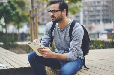Konsantre yetenekli sakallı ressam ile dış görev ve çizim şık optik gözlük çantası sokakta oturan Not Defteri'nde skeçler. Dalgın yetenekli sanatçı kopya kentsel ayarı not defterinde