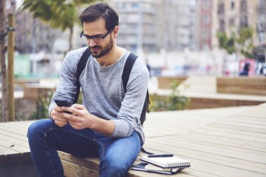 Kentsel ayarlarında oturan modern telefon e-posta yazma gözlük dalgın sakallı yolcu. Sırt çantası yüksek olarak bağlanan cep telefonunda sohbet ile erkek öğrenci hızda internet
