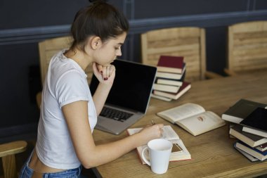 Dalgın öğrenci kitap okuma ve kitaplık modern macbook bilgisayar ile oturup sınava hazırlık sırasında Notlar yapma. Bilgi öğrenme ve bazı kayıtlarını yazma akıllı kadın konsantre