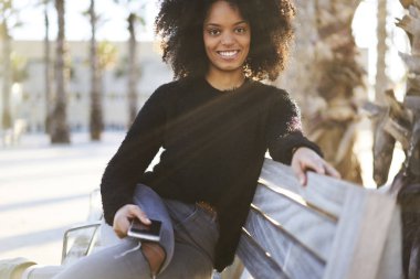 Yarım uzunluk portre sevimli pozitif kadın tezgah açık havada oturma ve modern mermiyi tutuyor kameraya bakarak gündelik kıyafet giymiş. Ücretsiz vaktinizi kıvırcık saçlı güzel kadın