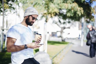 Yakışıklı erkek hipster dinlenme trendy güneş gözlüğü 4 g internet açık havada için bağlı smartphone video izlerken İspanyol modeli bloglama web sitesinde gidip hava bahar kahve zevk sakallı