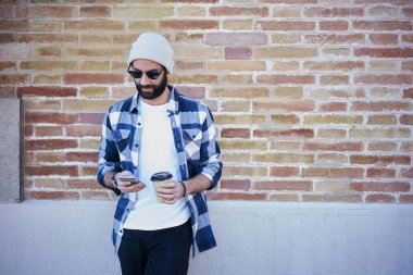 Şık giyimli hipster adam akıllı 4 g internet bağlantısı duvar promosyon arka plan, erkek üzerinde duran sakallı turist ile kahve açık havada haber okumak dinlenme kullanarak uygulama yükleme