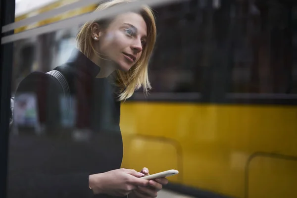 魅力的な金髪の女性観光客バス停市に移動するためのアプリケーションを使用しての公共交通機関を待っている魅力的なスマート フォンで 市街でのトラフィックの情報を閲覧する内気な少女 — ストック写真