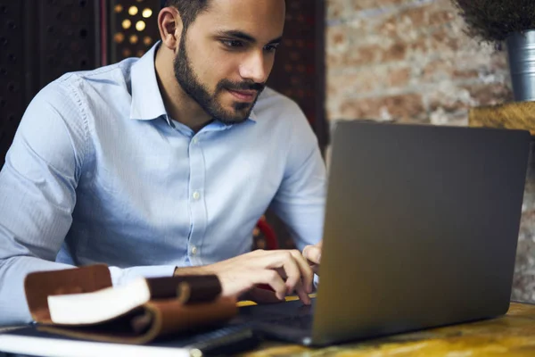 Concentrated handsome male businessman working via laptop computer checking email box waiting for receiving report, pensive freelancer creating advertising content earning money online sitting in cafe