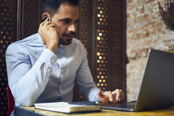 Handsome skilled copywriter discussing ideas for working task having phone talk sitting near laptop computer connected to wifi in cafe, confident young freelancer confirming booking calling to service