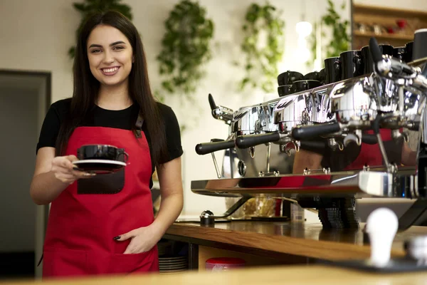 Portrait of smiling young business woman working barista in stylish restaurant interior.Cheerful waitress holding aroma coffee in hand made in modern machine.Female entrepreneur in red apron looking at camera