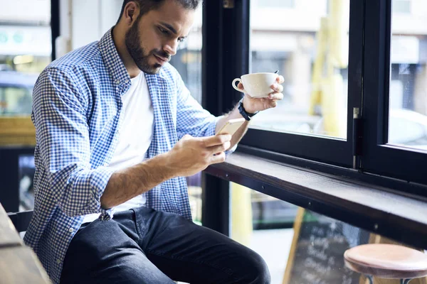 Handsome male blogger checking account in social networks updating information on profile suing mobile ,young casually dressed student reading news browsing information on networks during coffee break