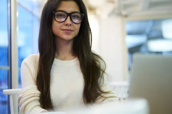 Confident brunette female employee of company looking at camera while working in office sitting near blurred advertising area, portrait of confident manager in trendy eyewear concentrated on job