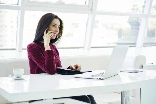 ラップトップ コンピューターで作業中に携帯電話で友人と面白い会話を持つ陽気な女性 笑顔マネージャー オフィスで近代的な携帯電話を介してクライアントと通信します コピー スペース — ストック写真