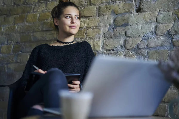 Attractive young author looking away while writing notes in notepad sitting in coworking space at laptop computer.Beautiful hipster girl thinking on plans and recording checklist in notebook